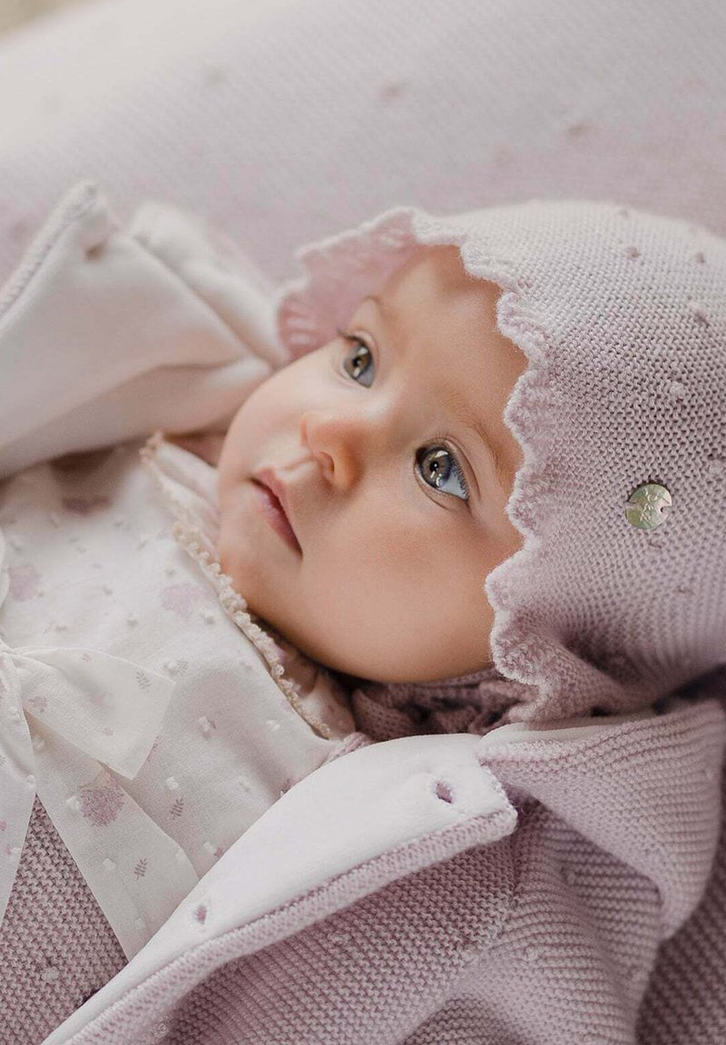 Bébé portant un doux bonnet tricoté rose avec des bords festonnés, une robe claire ornée de motifs floraux et de détails en nœud, blotti dans un tissu douillet.