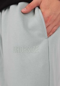 Világoskék, puha anyagból készült melegítők, bordázott derékszalaggal. A "DROPSIZE" felirat finoman, fényes kivitelben van nyomtatva az elülső zseb területén.