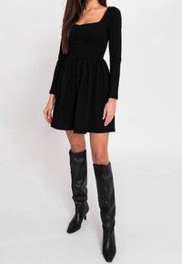 Femme portant une robe noire à manches longues avec un décolleté carré et des bottes en cuir noires montantes jusqu'aux genoux, debout devant un fond uni.