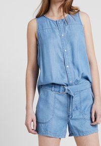 Ljust blå denim utan ärmar byxdress med knäppning framtill, bälte i midjan och framfickor, med en avslappnad passform och slät textur.