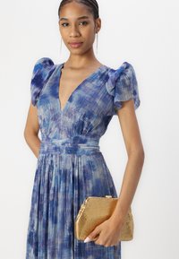 Pleated, V-kauluksen mekko sinisen ja violetin sävyissä, puffihäntäiset hihat. Kulta metallinen laukku, jossa on ruudullinen kuvio, pidetty kädessä.