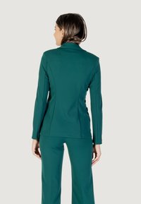 Una mujer con un traje verde se encuentra de espaldas, mostrando una chaqueta ajustada y pantalones a juego sobre un fondo neutro.