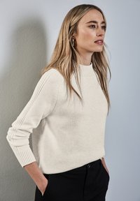 Crème gebreide sweater met een ribbelrond decolleté, ribbelmanchetten en verticale stikdetails langs de mouwen. Draag met zwarte broek.