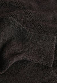 Tmavá vlnená tkanina s jemnými vzormi, zložená tak, aby ukázala hladký a teplý povrch. Obsahuje manžetu s rebrovaným detailom.