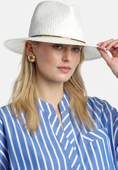Femme blonde portant de grandes boucles d'oreilles en or, un chapeau blanc texturé à large bord, et une chemise rayée bleue et blanche, tenant le bord du chapeau.