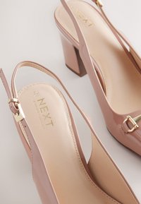 Paire d'escarpins nus en cuir verni avec talons blocs et détail de boucle dorée sur fond blanc.