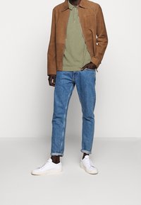 Hnědá semišová bunda přes zelené polo tričko, doplněná světle modrými džíny a bílými teniskami. Oblečení má ležérní, uvolněný střih.