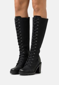 Bottes hautes noires jusqu'au genou en matériau texturé, présentant un design à lacets, des œillets argentés et un talon épais avec une semelle crantée.
