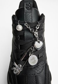 Zwarte sneaker met zwarte veters versierd met zilveren bedels waaronder een parel, hartje, rond logo en strik op een veiligheidsspeld.