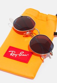 Ray-Ban saulės akiniai su tamsiais lęšiais, sidabro metaliniu rėmeliu ir skaidriais nosies pagalvėlėmis, ilsisi ant ryškių oranžinių audinių maišelių su raudonu logotipu.