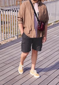 Camisa de botones marrón sobre una camiseta ligera púrpura, pantalones cortos de cargo negros y zapatillas beige. Se lleva un pequeño bolso cruzado burdeos.