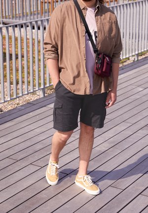 Chemise marron à boutons sur un t-shirt lilas clair, shorts cargo noirs et baskets beiges. Un petit sac bandoulière bordeaux est porté.