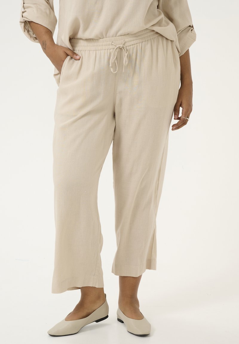 Pantalones de lino beige con cintura elástica y cordón. Presentan un largo recortado y bolsillos laterales. Se llevan con zapatos planos ligeros.