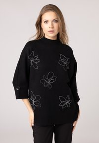 Maglione nero con colletto alto, vestibilità over, e maniche 3/4. Presenta ricami floreali bianchi e dettagli a bottone sulle maniche.