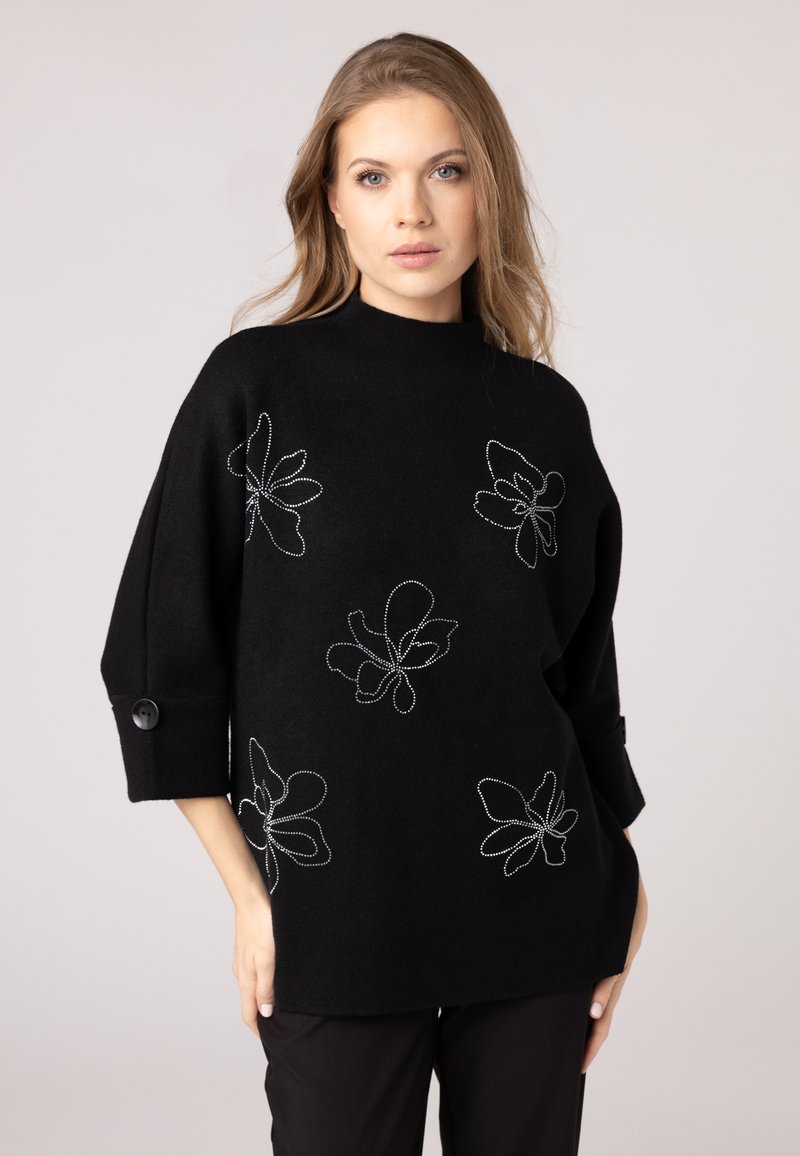 Maglione nero con colletto alto, vestibilità over, e maniche 3/4. Presenta ricami floreali bianchi e dettagli a bottone sulle maniche.
