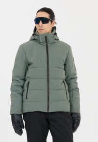 Giacca imbottita con zip in verde tenue, dotata di cappuccio e con dettagli in cerniera nera e tasche. Il modello indossa guanti neri e occhiali da sole.