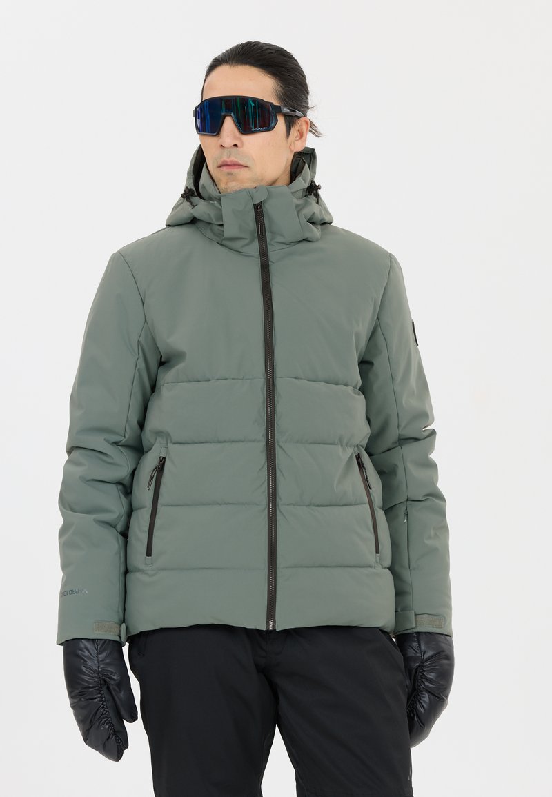 Gepolsterte, grünliche Jacke mit Reißverschluss und Kapuze, mit schwarzen Reißverschlussakzenten und Taschen. Das Modell trägt schwarze Handschuhe und Sonnenbrille.