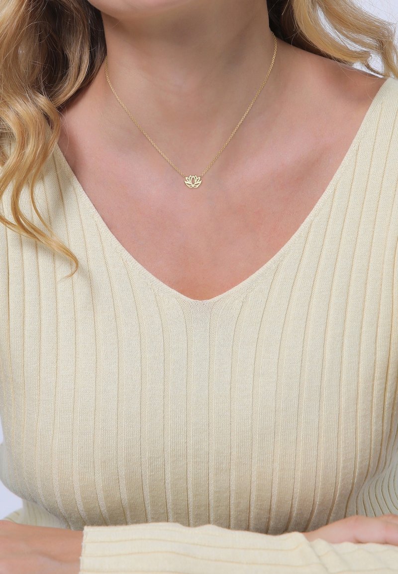 Femme portant un collier en or avec une fleur de lotus et une fine chaîne, ainsi qu'un pull crème à côtes avec un col en V, les bras croisés sous la poitrine.