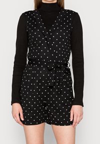 Svart prickig klänning med omslagdesign, knytmidja och korta byxor. Lager på en svart polotröja, vilket ger en texturerad look.