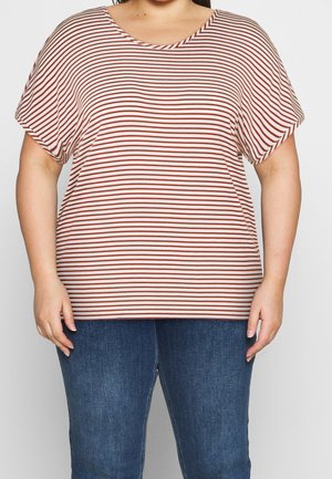 Femme portant une chemise ample à manches courtes avec des rayures horizontales rouges et blanches et un jean bleu, debout devant un fond uni.