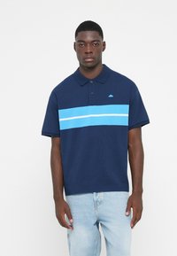 Marininspirerad polo skjorta med korta ärmar, med horisontella ljusblå och vita ränder. Inkluderar en liten logotyp på vänster bröst. Tillverkad av bomull.