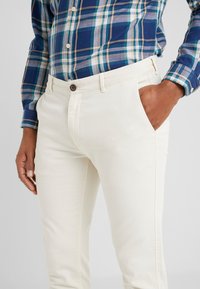 Pantalon beige en coton à coupe ajustée, avec poches latérales et fermeture par bouton marron. Porté avec une chemise à carreaux bleue et verte.