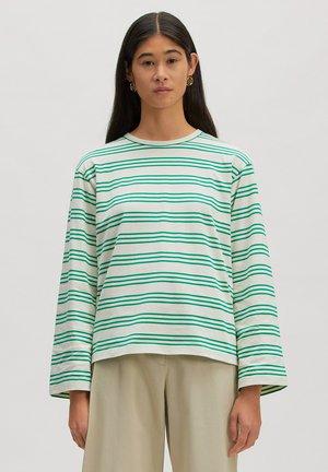 Femme aux longs cheveux foncés portant des boucles d'oreilles créoles dorées, une chemise blanche à manches longues avec des rayures horizontales vertes et un pantalon beige, debout devant un fond uni.