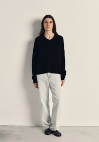 Črn pleten kardigan s okroglo ovratnikom, z gumbi spredaj, v kombinaciji s svetlobluzimi skinny hlačami in črnimi tkanimi sandalami.