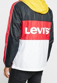 Osoba ubrana w kurtkę Levi's w blokach kolorów: czarna, czerwona, biała, z żółtym kapturem, pokazana od tyłu i z boku.