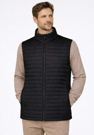Uomo che indossa un gilet imbottito nero senza maniche sopra una camicia beige a maniche lunghe, con le mani nelle tasche del gilet, guarda verso destra.