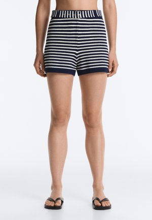Kvinde iført marineblå og hvide horisontale stribede strikkede shorts med bælte, parret med sorte flipflops, stående mod en hvid baggrund.