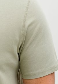 Κοντινό πλάνο μανικιού και ώμου ενός ανοιχτοπράσινου κοντομάνικου T-shirt που δείχνει την υφή του υφάσματος και τη ραφή στο μπράτσο ενός ατόμου.