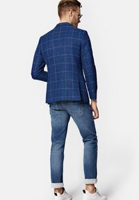 Niebieska marynarka w kratę z kontrastowymi liniami, noszona na jasnych dżinsach. Ubranie ma podwinięte mankiety i białe trampki.