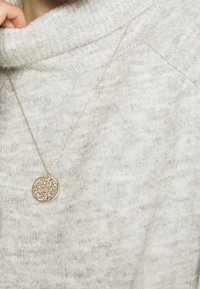Collier ton or avec un pendentif circulaire finement texturé, porté sur un pull à col roulé en tricot gris clair.