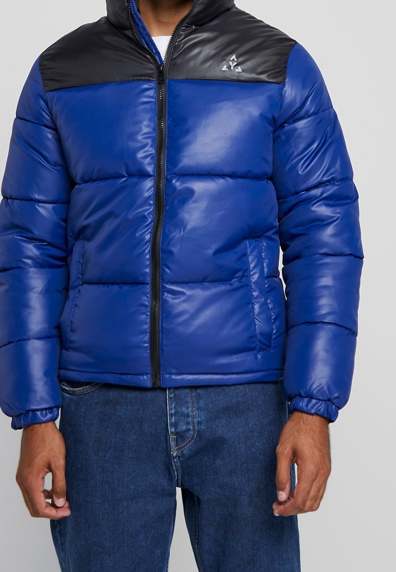 Blå puffjacka med en svart övre panel, utrustad med en framsidans blixtlås, elastiska ärmslut och vertikalt quiltat mönster. Accenterad med en logotyp på bröstet.