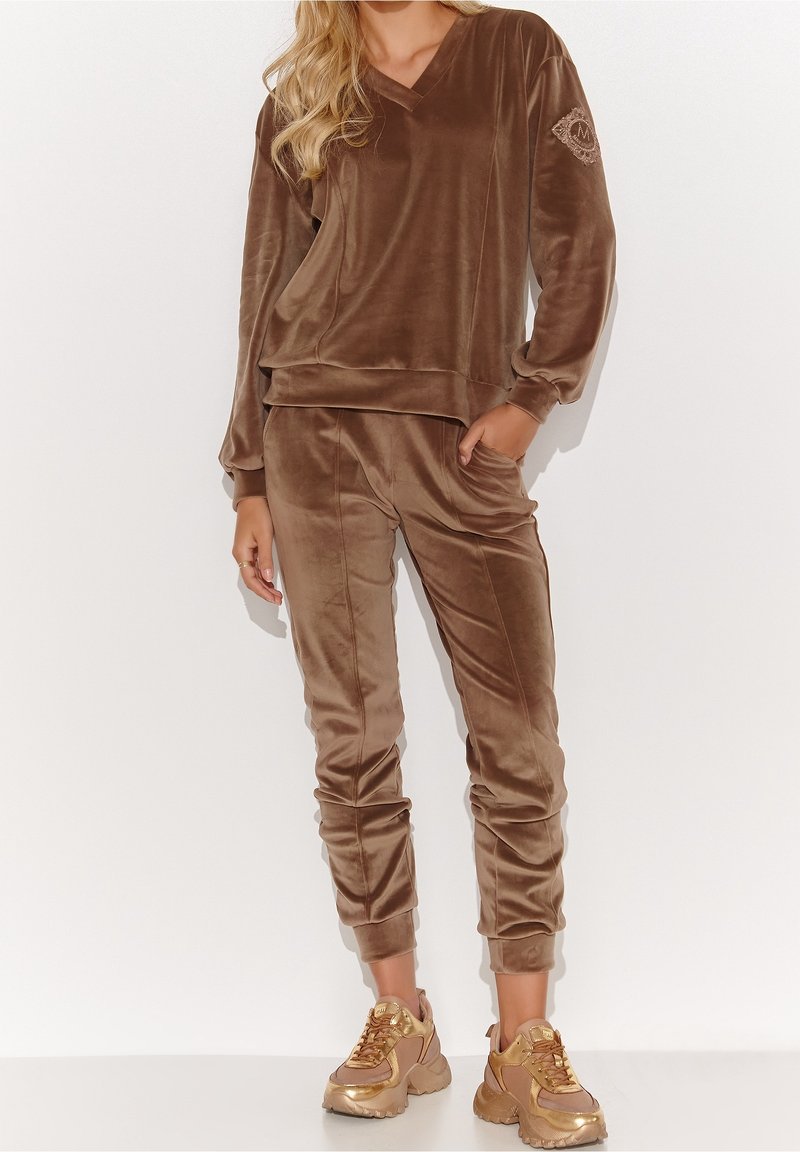Femme portant un survêtement marron en velours avec manches longues et pantalon de jogging assorti, associé à des baskets épaisses dorées, debout sur fond blanc.