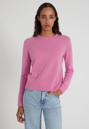 Donna con capelli ricci che indossa un maglione di lana rosa acceso e jeans azzurri, in piedi con una mano in tasca su uno sfondo uniforme.