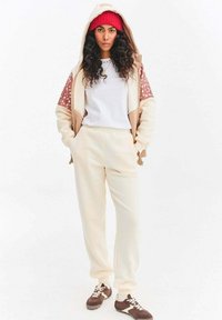 Kremowe spodnie dresowe i bluza na suwak z wzorzystymi rękawami, noszone na białej koszulce; do tego brązowo-białe trampki.