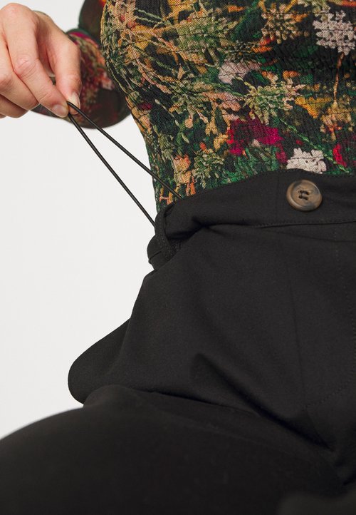 Czarne spodnie z wysokim stanem, zapinane na guzik, z regulowanym sznurkiem. Teksturowana, kolorowa bluzka z wzorem częściowo widoczna nad talią.