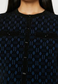 Cardigan boutonné à motifs noirs et bleus avec col rond et deux poches poitrine sur une personne aux longs cheveux foncés.
