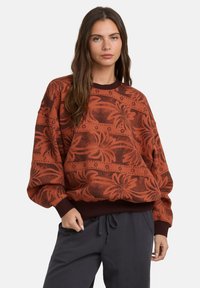 Czerwony sweter z długim rękawem w wzory, z kwiatowym motywem oraz ściągaczami przy mankietach i dekolcie. W połączeniu z ciemnoszarymi spodniami.