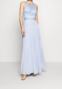 Světle modré večerní šaty až na zem s krajkovým korpusem se složitými květinovými vzory, obepnutým pasem a splývavou sukní. Celkový dojem dotvářejí jednoduché nahé sandály na podpatku.