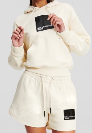 Personne portant un sweat à capuche crème et un short assorti avec des logos carrés noirs indiquant "Karl Lagerfeld Jeans" sur la poitrine et la jambe.