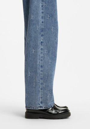 Jean en denim bleu avec motif script blanc répété, porté avec des mocassins en cuir noir, montré de profil sur un fond uni.