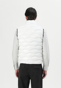 White puffer vest with quilted, wave-like pattern, high collar, and side zip details, worn over a striped long-sleeve shirt. Black trousers.