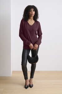 Bordeauxfarbens Langarmshirt mit V-Ausschnitt, kombiniert mit schwarzen engen Hosen aus Kunstleder und einer schwarzen Handtasche in Halbmondform. Spitz zulaufende schwarze Schuhe.