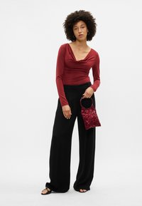 Burgundska bluza s dolgimi rokavi in drapirano ovratnikom, v kombinaciji s črnimi širokimi hlačami in majhno burguindkasto torbico s bleščicami.