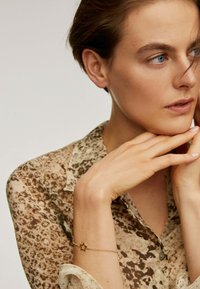 Blusa marrón transparente con un patrón moteado; pulsera de estrellas doradas en la muñeca; manos tocando suavemente la cara; suave iluminación natural.