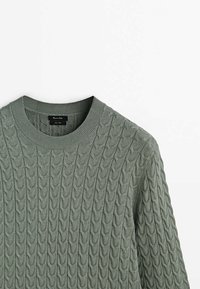 Vihreä palmikkoneulecrewneck-paita, jossa on ribbireunus, valmistettu teksturoidusta kankaasta, aseteltu tasaisesti valkoista taustaa vasten.