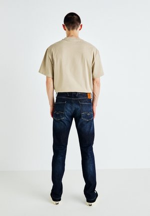 Mand iført beige kortærmet skjorte, mørkeblå jeans med baglommer og sorte og hvide sko, der står med front mod en hvid væg.